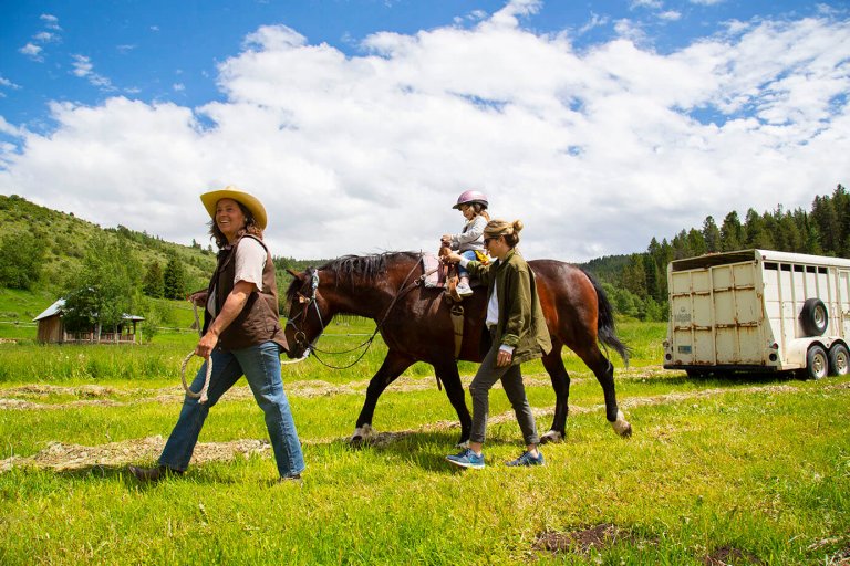 A child riding a horse being led by adult as another adult walks beside her in a wide-open field of grass surrounded by trees.