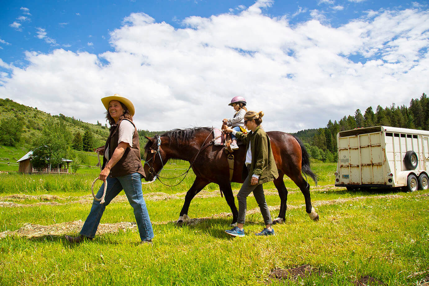 A child riding a horse being led by adult as another adult walks beside her in a wide-open field of grass surrounded by trees.