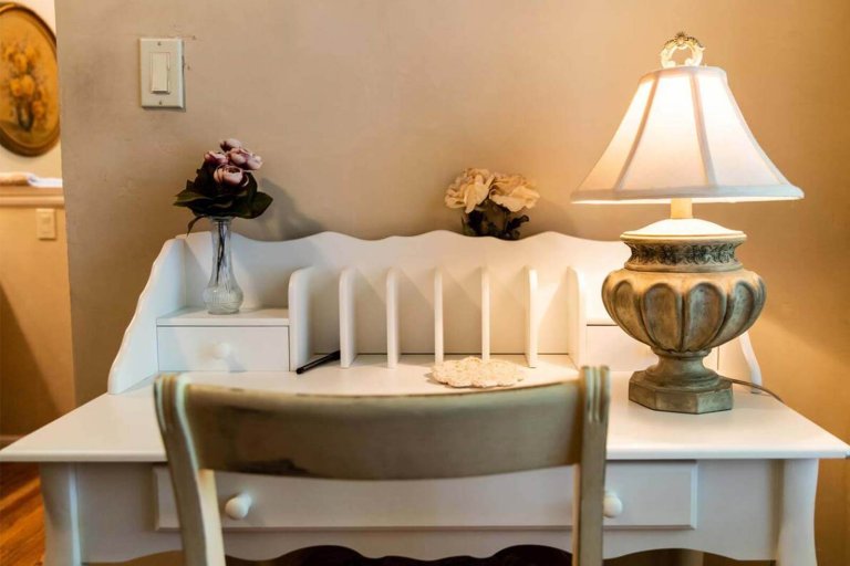 A chair in front of a small white desk with vases of flowers and a table lamp.