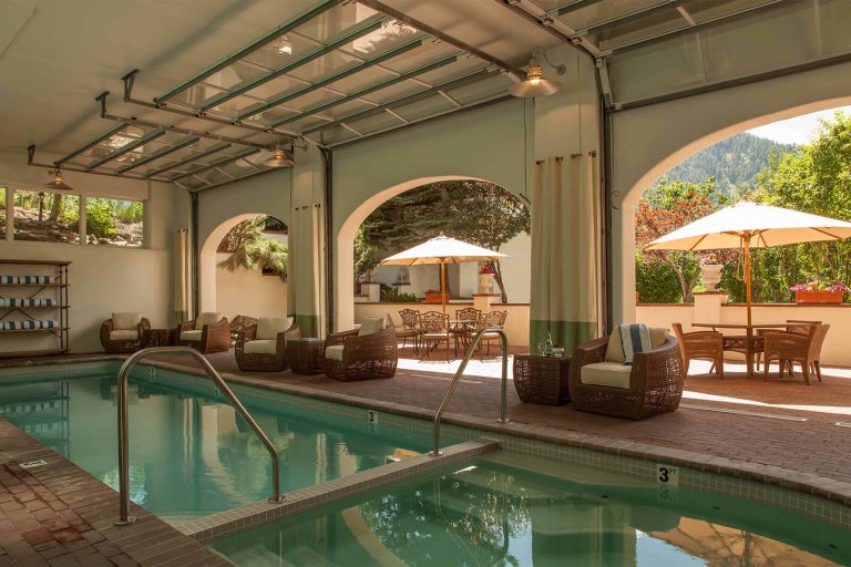 An indoor hotel pool lined with wicker chairs, and through the doors just outside, two tables with wicker chairs and umbrellas.