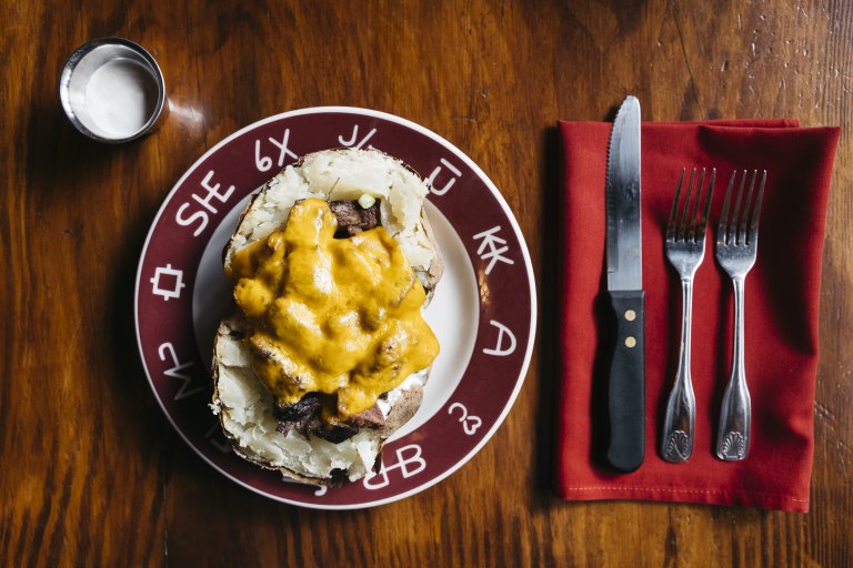 A top view of a large baked potato topped with beef and melted cheese on a plate between a cup of sour cream and a knife and two forks on a red cloth napkin on a table at the Pioneer Saloon.