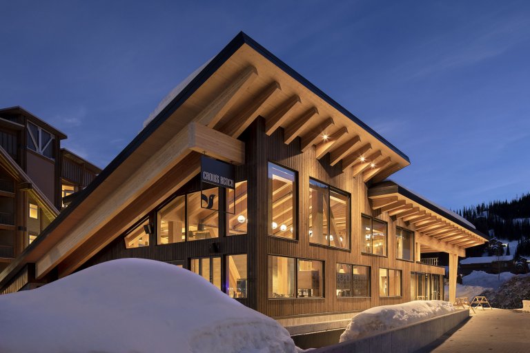 An angular ski lodge with large windows and another building behind it surrounded by piles of shoveled snow and trees.
