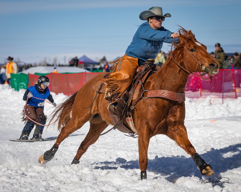 A person riding a galloping horse through a field of snow pulling a person riding a snowboard.