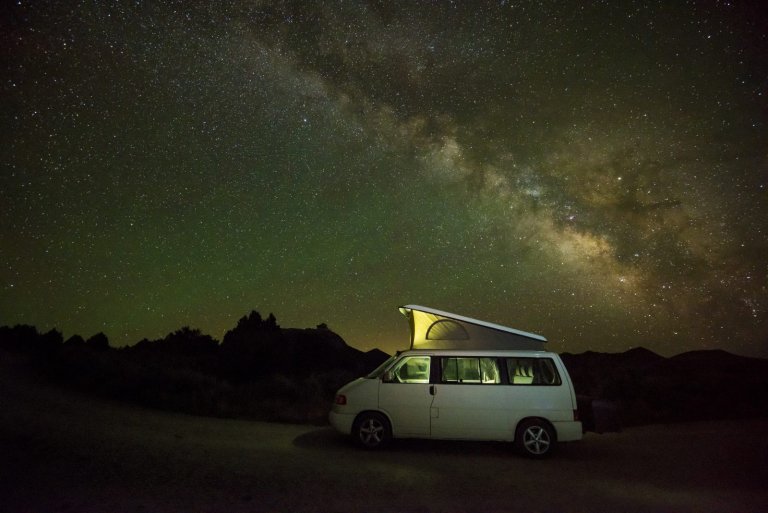 A camper van with a tent on the roof is parked at night with the Milky Way visible in the sky above it.